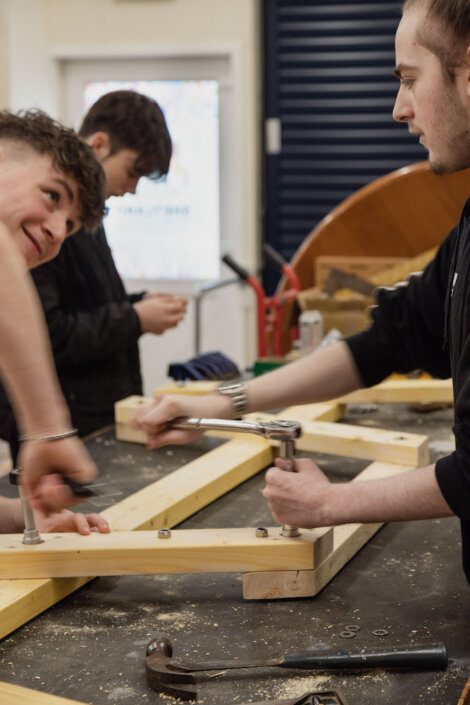 Three people working on a wooden structure in a workshop, using tools such as a wrench and crowbar.