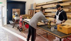 Two individuals work on a carpentry project, using drills and other tools, in a well-lit workshop with shelves of wood in the background.