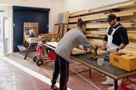 Two individuals work on a carpentry project, using drills and other tools, in a well-lit workshop with shelves of wood in the background.