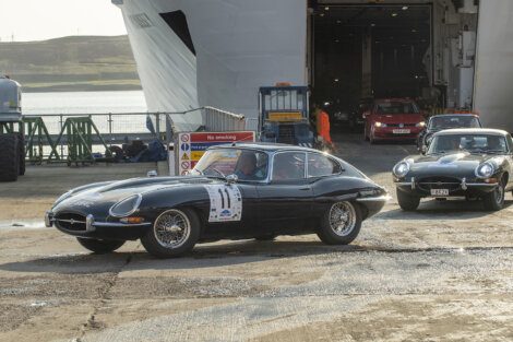 Two vintage sports cars drive off a ferry. The lead car has a racing number on the door, with another similar car following behind. The background shows the open ferry ramp and industrial surroundings.