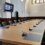 A conference room with a long wooden table and multiple chairs. Microphones are set up at each seat. A video conference is displayed on a wall-mounted screen.