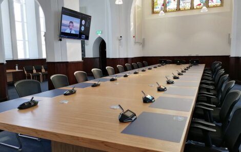 A conference room with a long wooden table and multiple chairs. Microphones are set up at each seat. A video conference is displayed on a wall-mounted screen.
