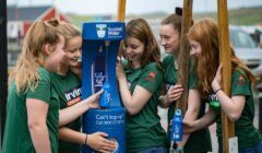 Five young women in green shirts fill blue water bottles at a Scottish Water refill station outdoors.