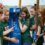 Five young women in green shirts fill blue water bottles at a Scottish Water refill station outdoors.