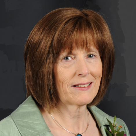 A middle-aged woman with shoulder-length brown hair and bangs, wearing a light green jacket and a necklace, poses against a dark background.