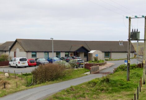 A single-story building with a slanted roof, several parked cars in front, and some greenery around it. A small road and utility pole are visible in the foreground.