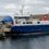 A blue and white ferry named "Linga" docks at a rocky pier in a calm coastal area under a cloudy sky.