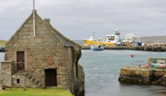 Old stone building by the sea with several boats docked at a nearby pier. Overcast sky.