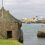 Old stone building by the sea with several boats docked at a nearby pier. Overcast sky.