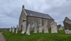 A small stone church with several old, weathered gravestones in front, set against a cloudy sky.