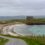 A narrow road leads to a stone house near a rocky shoreline with turquoise water and grassy landscape under a cloudy sky.
