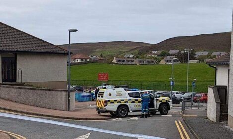 A police vehicle and officers are present at the entrance of a residential area with houses and hills in the background. A "School Patrol" sign is visible on the sidewalk.