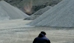 A person in dark clothing stands near large piles of gravel in an industrial area with a hill in the background.