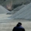 A person in dark clothing stands near large piles of gravel in an industrial area with a hill in the background.