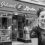 A woman stands smiling in front of a store named "Island Larder" which offers coffee, tea, hot chocolate, Shetland fudge, hampers, gifts, and other goods.