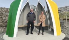 Two people stand under a colorful, flower-petal-shaped shelter with green, yellow, and white panels. Rocks and a stone wall are in the background, with a building visible in the distance.