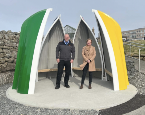 Two people stand under a colorful, flower-petal-shaped shelter with green, yellow, and white panels. Rocks and a stone wall are in the background, with a building visible in the distance.