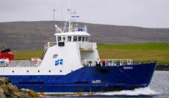 A blue and white ship named "BIGGA" is sailing near a rocky shore with hills in the background. There is a wind turbine visible on the hill.
