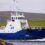 A blue and white ship named "BIGGA" is sailing near a rocky shore with hills in the background. There is a wind turbine visible on the hill.
