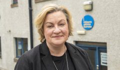 A person with short blonde hair and a black blazer stands outside a building with a sign that reads "Citizens Advice Bureau.