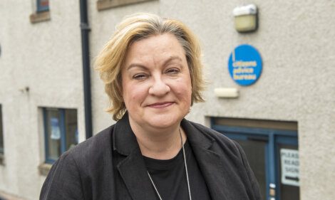 A person with short blonde hair and a black blazer stands outside a building with a sign that reads "Citizens Advice Bureau.