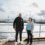 Two people stand in front of a harbor with a docked ship and buildings in the background on a cloudy day.