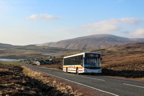 A bus marked "Toft 23" travels on a rural road surrounded by hills and scattered buildings under a clear sky.