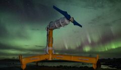 A tidal turbine stands against a backdrop of green aurora borealis in a dark night sky.