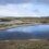 A calm lake reflecting the cloudy sky, surrounded by a barren, rocky landscape with sparse vegetation and distant hills.
