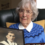 An elderly woman with white hair and glasses sits holding a framed black-and-white photograph of a man in graduation attire. She is wearing a blue and white sweater and is seated in a green armchair.