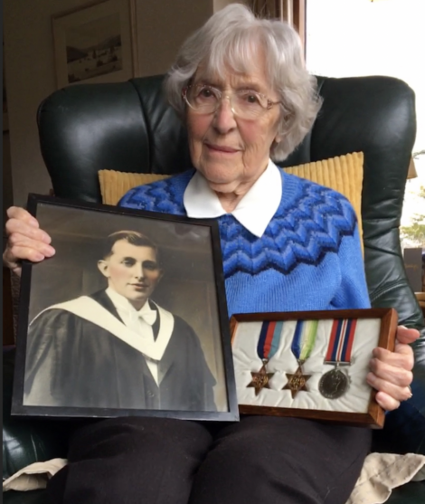 An elderly woman seated in a green armchair holds a framed photo of a man in academic attire and a display case with four military medals.