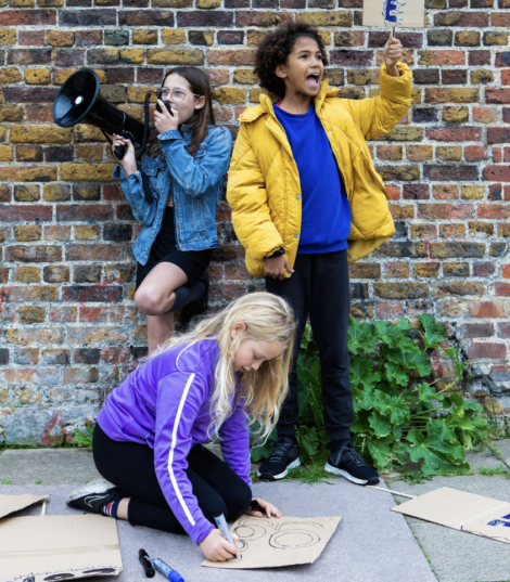 Three children engaged in a protest: one kneeling and drawing a sign, one holding a megaphone, and one wearing a yellow jacket, holding up a sign. They are in front of a brick wall with greenery at the base.