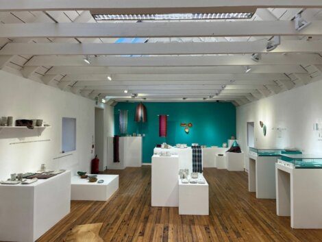 A well-lit gallery room showcases various artworks, including textiles and ceramic pieces, displayed on white pedestals and shelves with a teal accent wall in the background.