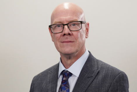 Bald man with glasses wearing a gray suit, light blue shirt, and patterned tie, standing against a plain background.