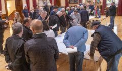 A group of people gather around tables in a hall, closely examining documents and objects on display. Some individuals are engaged in discussions while others are observing the materials.