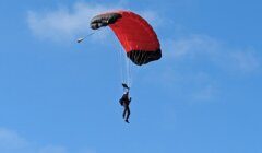 A person is mid-air parachuting with a red and black parachute against a clear blue sky.