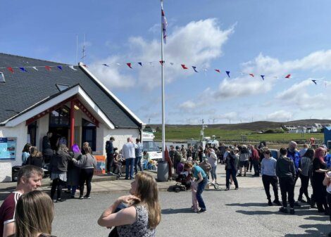 A crowd gathers outside a building with red trim and a flagpole. Bunting decorates the scene. The sky is partly cloudy, and hills are visible in the background.