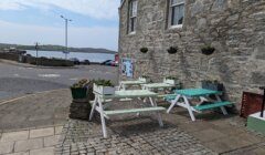 Outdoor seating area with two picnic tables next to a stone building, overlooking a parking lot and a view of the sea in the distance.