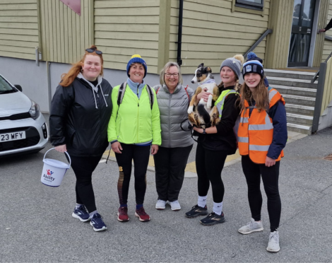 Five women, dressed in casual outdoor attire, stand together smiling for a photo. One woman is holding a dog. Another holds a collection bucket. They are outdoors near a building with steps.