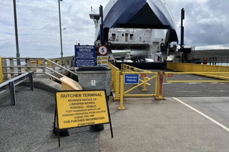 A ferry docked at Gutcher Terminal with a sign warning about upcoming linkspan works and alternative foot passenger arrangements. A passenger walkway and a customer notice are also visible.