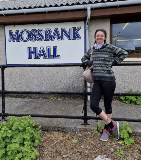 A person smiling and standing in front of a building with a sign that reads "MOSSBANK HALL," wearing a patterned sweater and black leggings.