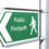 Green and white sign on a post indicating a public footpath with a pedestrian icon pointing left.