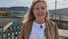 Woman standing outdoors smiling with mountains in the background and children's playground equipment visible behind a fence.