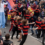 A group of people in matching red and black striped shirts dance in the street during a crowded parade. A person on stilts is visible in the background. Spectators line both sides of the street.