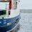 A ferry named "Fivla Lerwick" is docked at a harbor with land visible in the background. The ferry's hull is blue and white with red accents.