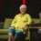 An elderly woman with short white hair, glasses, and a green shirt sits on a green sofa during an interview. She faces the interviewer, who holds papers and sits at a table with a glass of water.