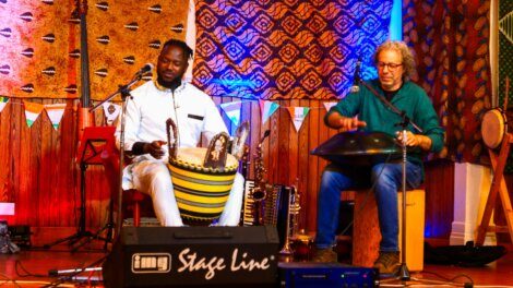 Two musicians perform on stage, one playing a traditional drum while the other plays a handpan. The backdrop features patterned tapestries, and various musical instruments are visible in the background.