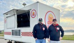 Two men wearing matching hoodies and caps stand in front of a white food truck with "Snack & Chat 2 Cows" logos on it. The food truck is parked outdoors with scenic landscape in the background.