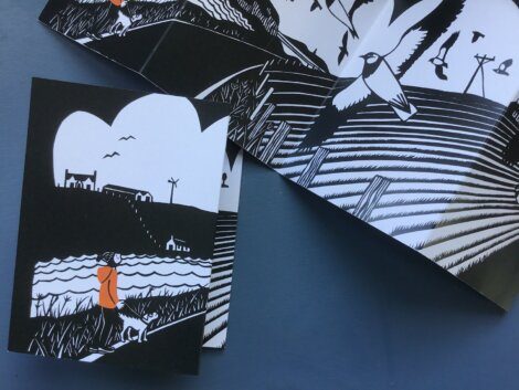 A folded black and white illustration booklet showing a person in an orange coat walking a dog by a lake, with birds, houses, and power lines in the background.