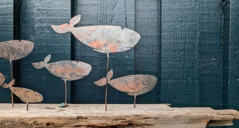Metal whale sculptures of various sizes mounted on thin rods are displayed on a wooden plank against a textured, dark blue wooden wall.